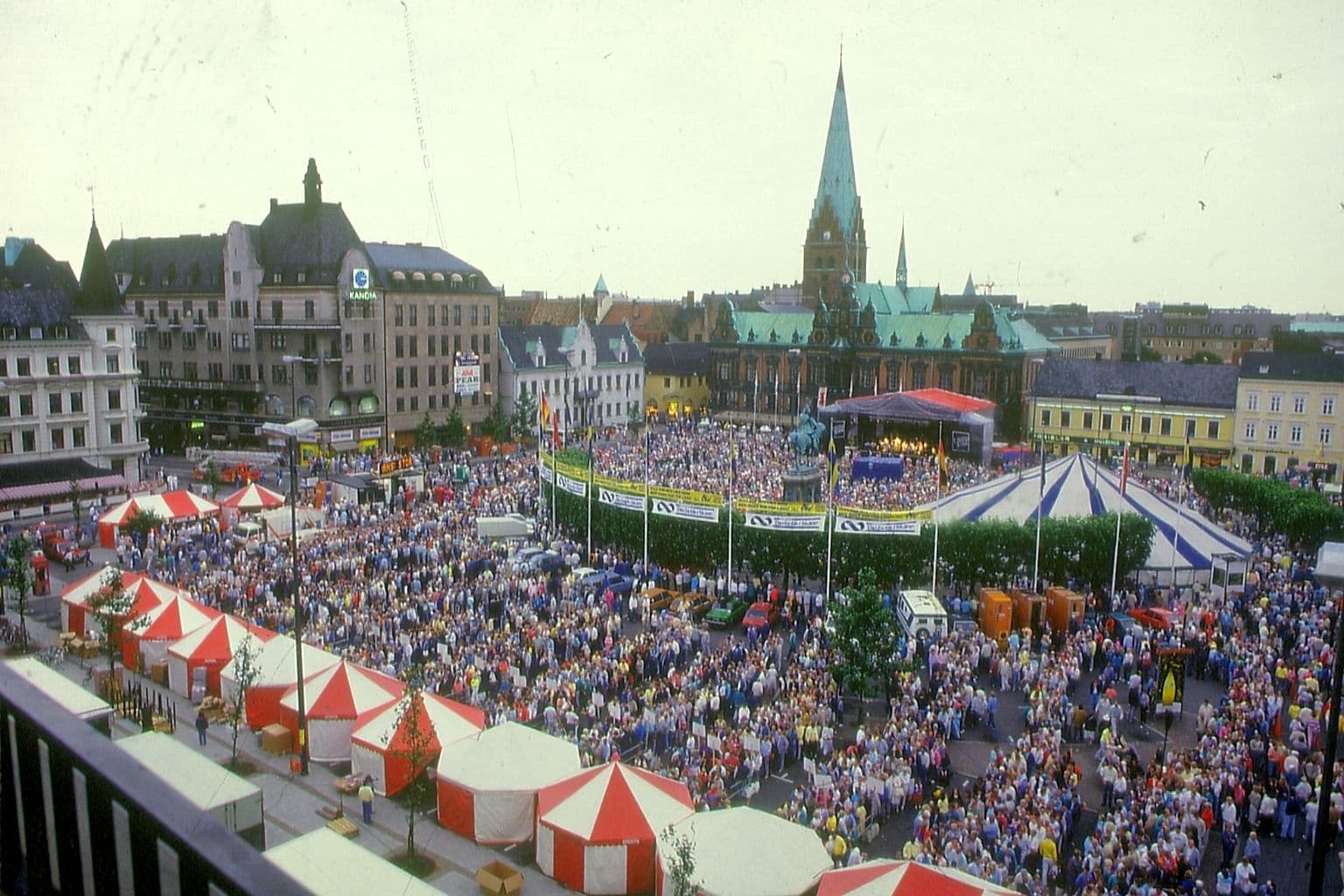 En flygbild över ett torg med en stor folkskara och några färgglada tält. I bakgrunden syns ett kyrktorn.