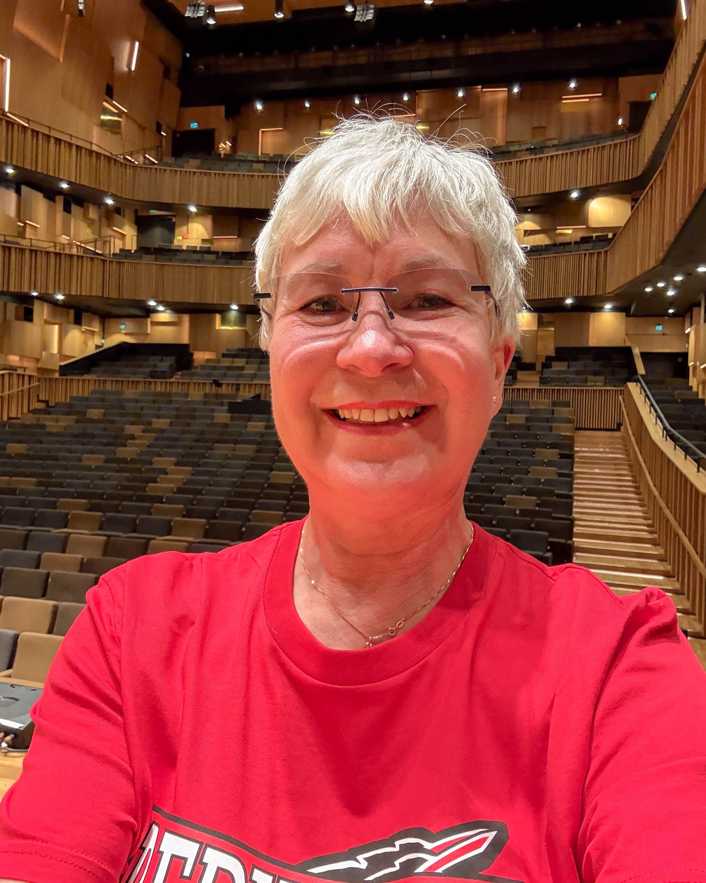 En äldre kvinna med glasögon i röd t-shirt. I bakgrunden syns konsertsalen på Malmö live.