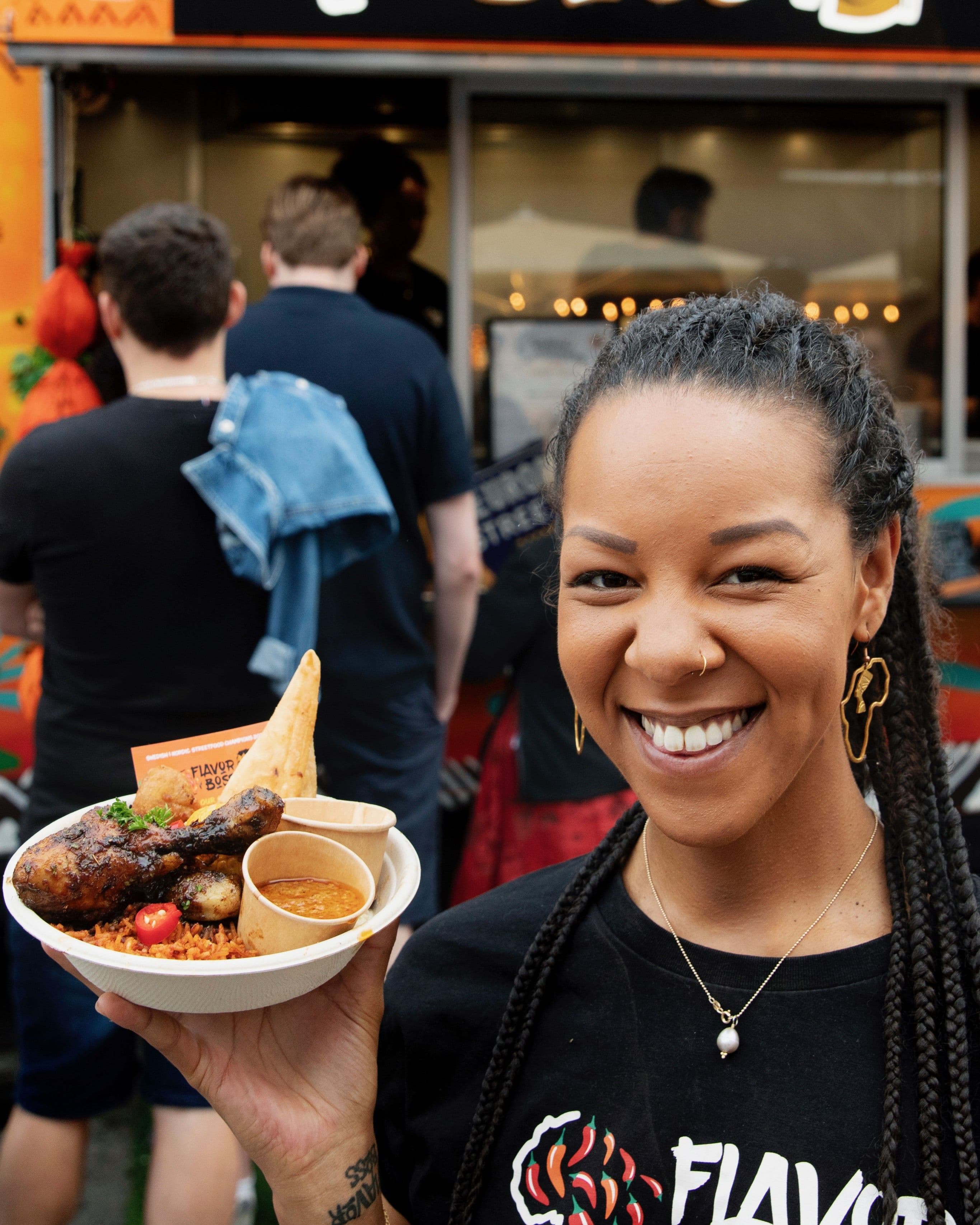 En kvinna i svart ti-shirt från med en tallrik med mat framför en food truck. Två män står i kö i bakgrunden.