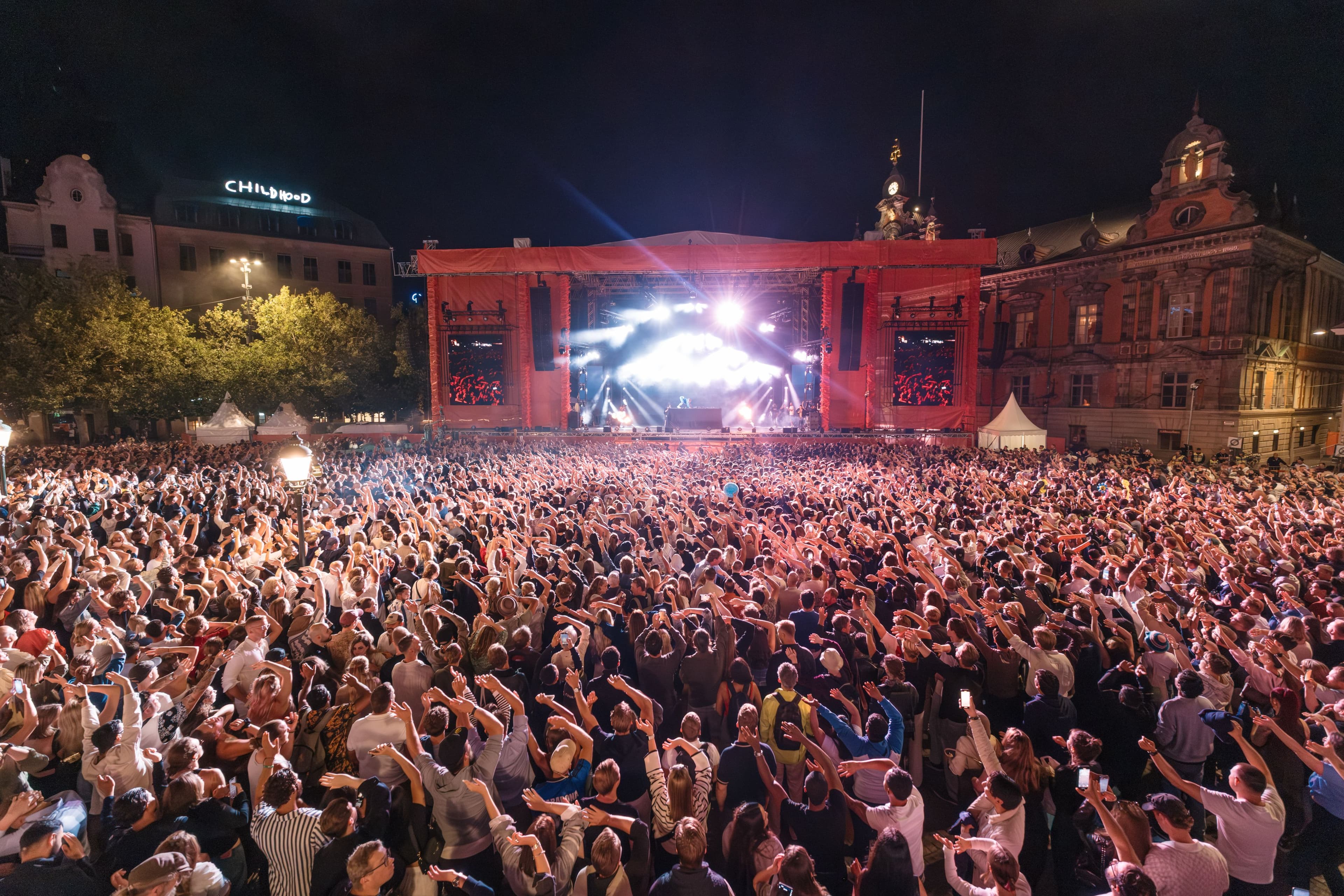 En dj uppträder på en stor scen på ett torg framför mycket publik som håller händerna i luften.