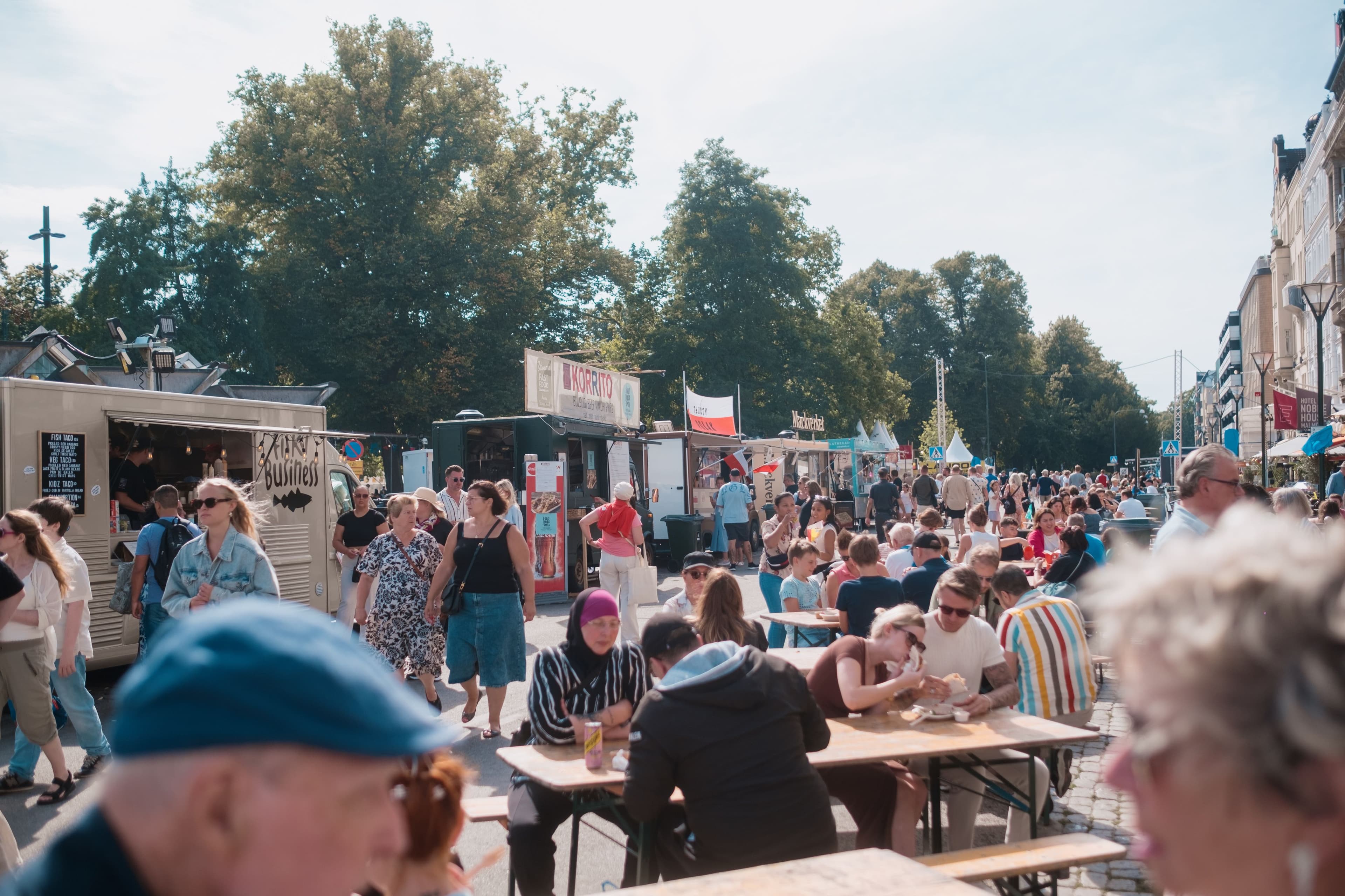 Besökare äter vid långbord och promenerar mellan food trucks på en solig gata under Malmöfestivalen.